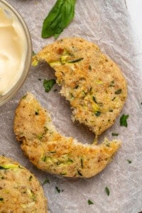 A close-up of a partially eaten zucchini fritter on parchment paper. The fritter is sprinkled with chopped herbs and paired with a small bowl of creamy dip. A basil leaf decorates the scene.