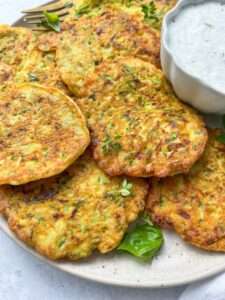 A plate of golden-brown zucchini fritters, made from a classic Zucchini Fritters Recipe, garnished with fresh herbs and served with a bowl of creamy dipping sauce.