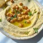 A bowl of creamy hummus from an easy hummus recipe is topped with whole chickpeas, a drizzle of olive oil, and fresh herbs, served with a tortilla chip on the side. The dish sits on a white surface in bright natural light.