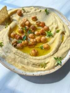 A bowl of creamy hummus from an easy hummus recipe is topped with whole chickpeas, a drizzle of olive oil, and fresh herbs, served with a tortilla chip on the side. The dish sits on a white surface in bright natural light.