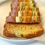 Several slices of moist Easy Zucchini Bread are arranged in a row on parchment paper, showcasing green zucchini pieces in the golden-brown loaf.