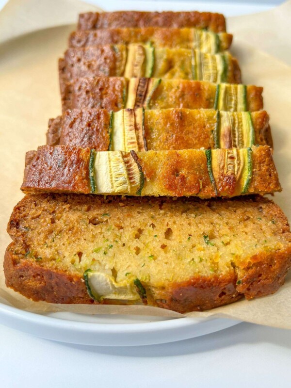 Several slices of moist Easy Zucchini Bread are arranged in a row on parchment paper, showcasing green zucchini pieces in the golden-brown loaf.