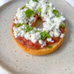 A toasted bagel half topped with tomato sauce, crumbled white cheese, and fresh basil leaves sits on a speckled beige plate—an irresistible twist on Greek Bruschetta.