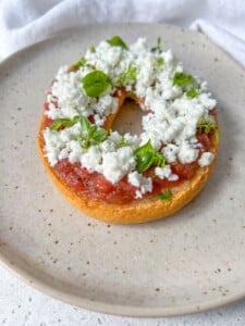 A toasted bagel half topped with tomato sauce, crumbled white cheese, and fresh basil leaves sits on a speckled beige plate—an irresistible twist on Greek Bruschetta.