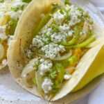 A close-up of a breakfast taco filled with sautéed onions, green peppers, crumbled white cheese, and fresh herbs on a light-colored plate.