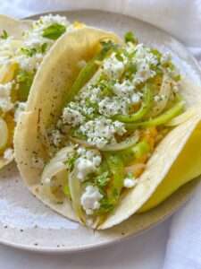 A close-up of a breakfast taco filled with sautéed onions, green peppers, crumbled white cheese, and fresh herbs on a light-colored plate.