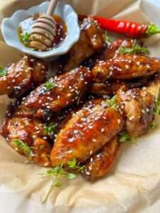 A plate of Honey BBQ Chicken Wings glazed to perfection, sprinkled with sesame seeds and fresh herbs, served with a small bowl of honey and a honey dipper, alongside a red chili pepper.