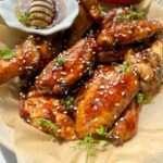 A plate of Honey BBQ Chicken Wings glazed to perfection, garnished with sesame seeds and fresh herbs, served alongside a small dish of honey with a dipper, and a vibrant red chili pepper on the side.