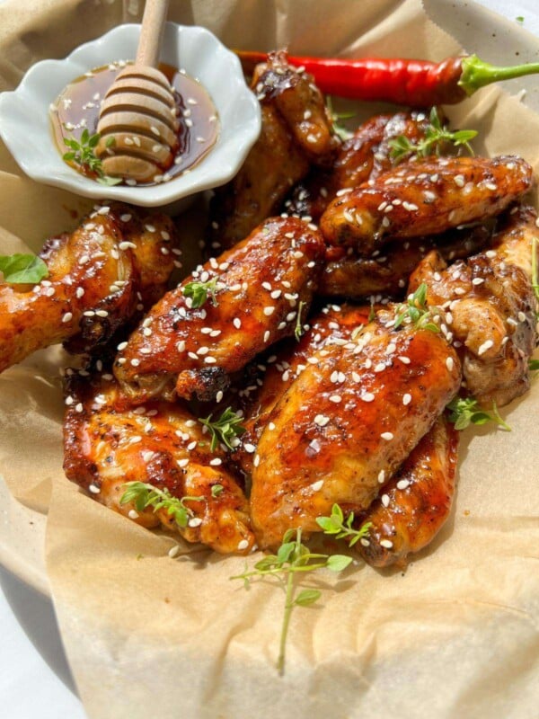 A plate of Honey BBQ Chicken Wings glazed to perfection, garnished with sesame seeds and fresh herbs, served alongside a small dish of honey with a dipper, and a vibrant red chili pepper on the side.