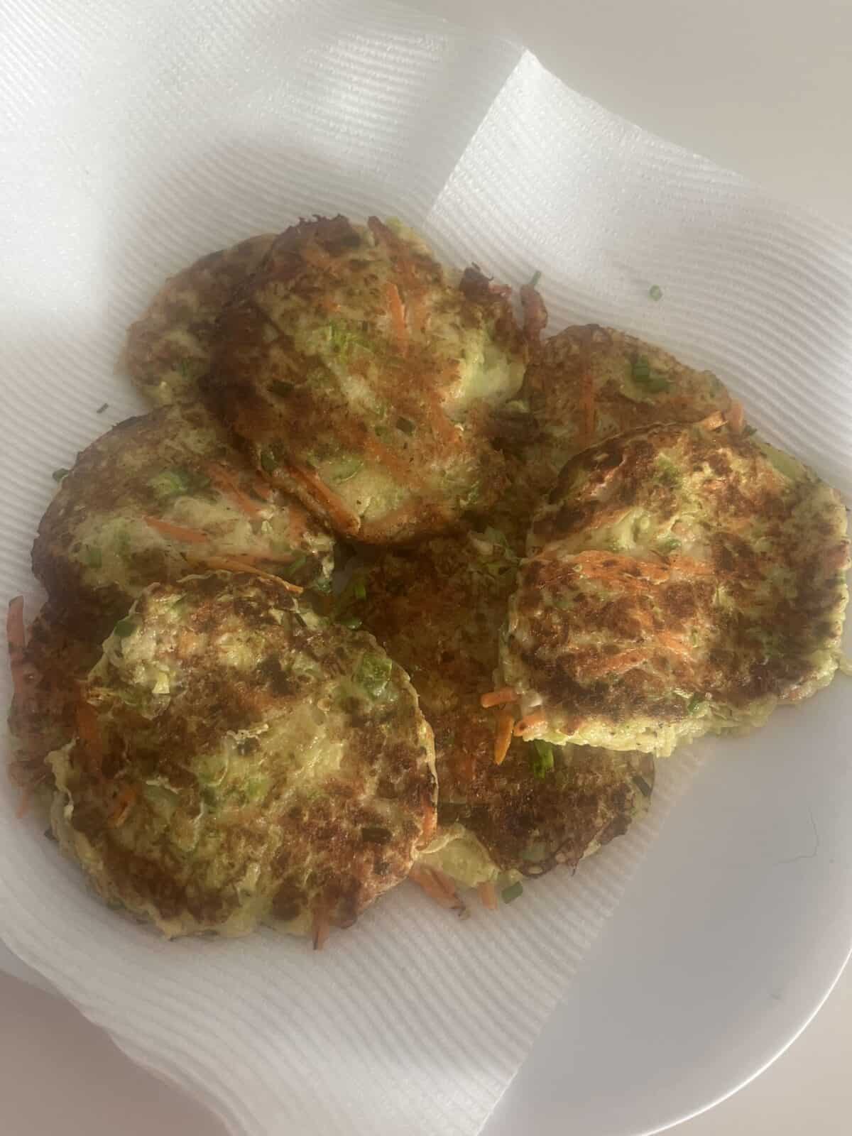 A plate of golden-brown vegetable fritters with visible pieces of carrot and green herbs, resting on a paper towel to absorb excess oil.