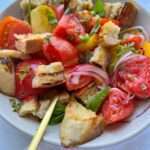A bowl of Panzanella Salad with colorful tomato wedges, toasted bread chunks, sliced red onions, basil leaves, and a light dressing is served with two gold forks.