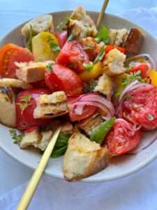 A bowl of Panzanella Salad with colorful tomato wedges, toasted bread chunks, sliced red onions, basil leaves, and a light dressing is served with two gold forks.