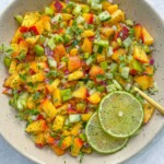 A bowl of vibrant Peach Salsa featuring diced cucumbers, red onions, yellow bell peppers, and peaches, garnished with fresh herbs and two slices of lime, sits on a light textured surface.