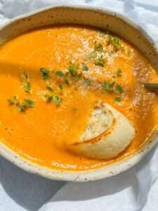 A bowl of creamy orange tomato soup, made from a delicious Creamy Tomato Soup Recipe, is garnished with fresh green herbs and served with a slice of crusty bread partially dipped in, bathed in bright natural light.