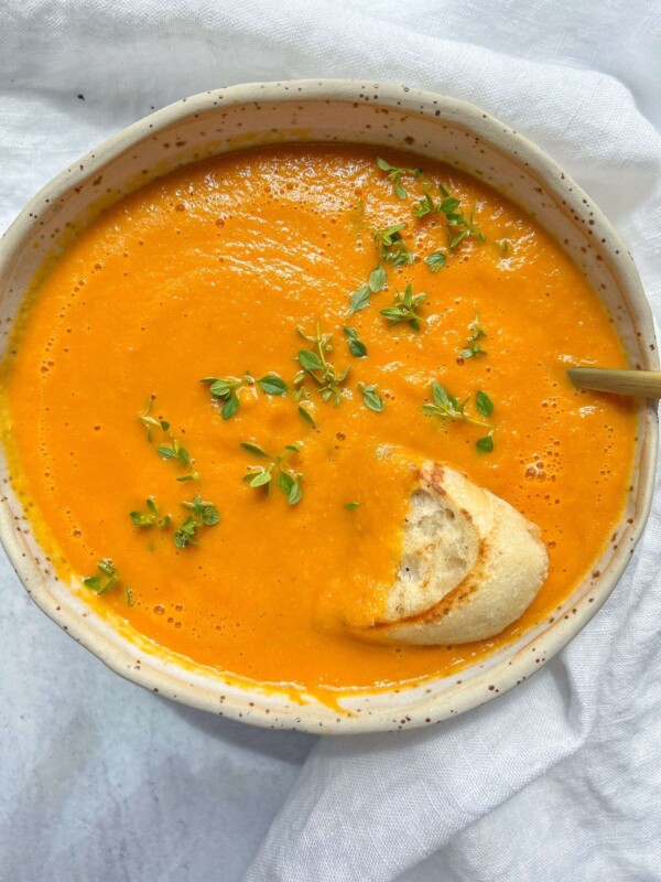 A bowl of creamy tomato soup recipe, vibrant orange and garnished with fresh green herbs, sits invitingly beside a piece of crusty bread partially dipped in the soup on a white cloth background.