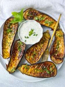 Roasted Eggplant Recipe: Roasted eggplant halves garnished with fresh herbs, arranged around a bowl of creamy white dipping sauce on a white plate, with a gold fork and basil sprigs for decoration.