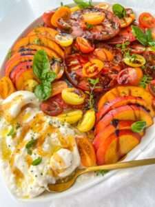 A colorful Burrata Tomato Peach Salad featuring sliced heirloom tomatoes, peaches, cherry tomatoes, fresh burrata cheese, basil, and herbs, drizzled with olive oil and balsamic glaze on a white plate with a gold spoon.