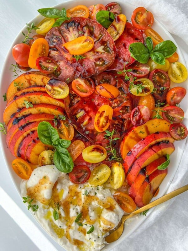 A vibrant Burrata Tomato Peach Salad with heirloom tomatoes, peaches, cherry tomatoes, fresh basil, and burrata cheese, drizzled with olive oil, served on a white plate with a gold fork.