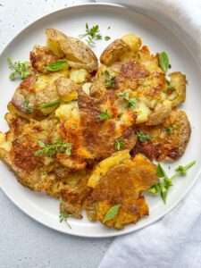 A white plate with golden, Crispy Smashed Potatoes garnished with fresh green herbs, set on a light-colored surface next to a white napkin.