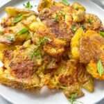A plate of Crispy Smashed Potatoes garnished with fresh green herbs, served on a white dish. The potatoes are golden brown with a slightly rustic texture, making them perfect for any crispy smashed potato lover.