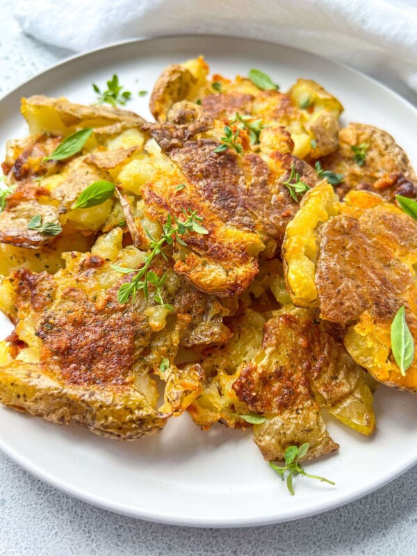 A plate of Crispy Smashed Potatoes garnished with fresh green herbs, served on a white dish. The potatoes are golden brown with a slightly rustic texture, making them perfect for any crispy smashed potato lover.