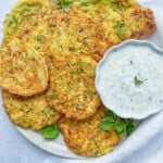 A plate of golden zucchini fritters from our favorite Zucchini Fritters Recipe, garnished with fresh herbs and served with creamy white dipping sauce, sits on a light background with a napkin and fork.