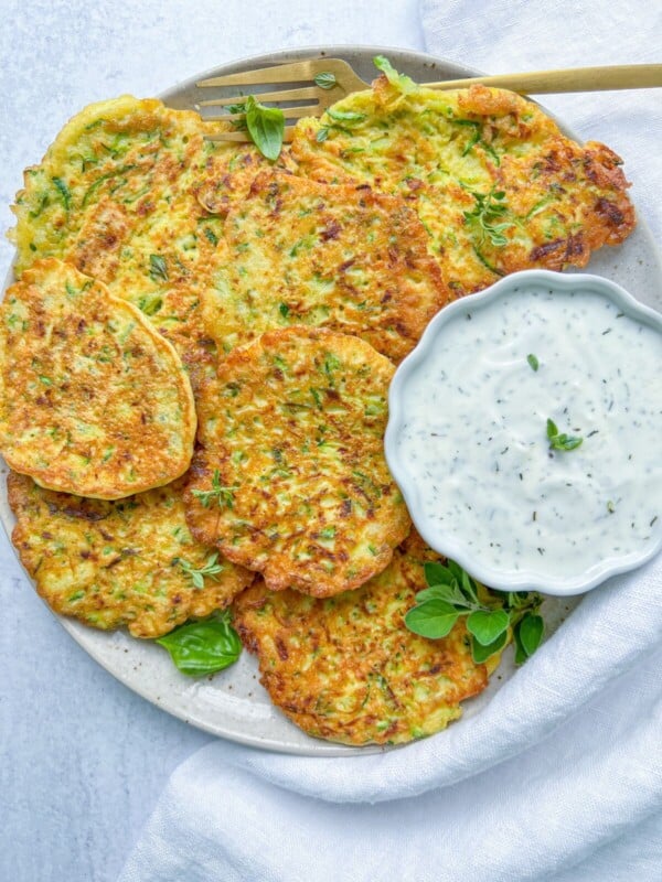 A plate of golden zucchini fritters from our favorite Zucchini Fritters Recipe, garnished with fresh herbs and served with creamy white dipping sauce, sits on a light background with a napkin and fork.