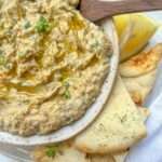 A bowl of creamy baba ghanoush, made from a classic Baba Ghanoush Recipe, topped with herbs and olive oil, served with pita bread slices and a lemon wedge on a plate. A wooden spoon rests on the bowl.