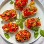 Six pieces of toasted bread topped with a colorful Italian Bruschetta mix are arranged on a white plate, garnished with fresh basil leaves and herbs.