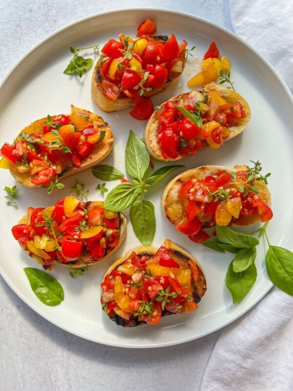 Six pieces of toasted bread topped with a colorful Italian Bruschetta mix are arranged on a white plate, garnished with fresh basil leaves and herbs.