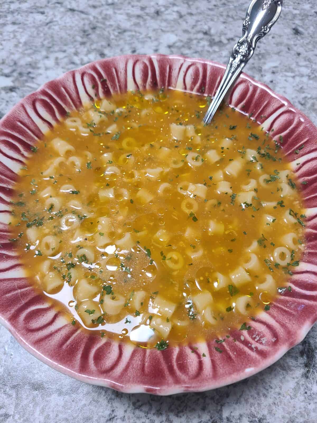 A pink-rimmed bowl filled with pasta soup featuring small ring-shaped noodles in a yellow broth, garnished with herbs, sits on a granite countertop with a silver spoon in the bowl.