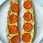 Two Zucchini Pizza Boats rest on a white plate, hollowed and filled with melted cheese, tomato sauce, and pepperoni slices, then garnished with fresh chopped chives.