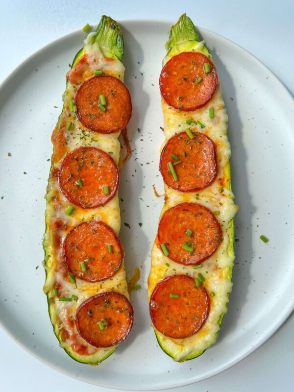 Two Zucchini Pizza Boats rest on a white plate, hollowed and filled with melted cheese, tomato sauce, and pepperoni slices, then garnished with fresh chopped chives.