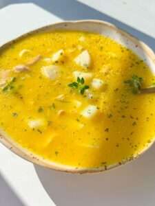 A bowl of Hidden Veggie Chicken Soup with chunks of potato, garnished with herbs, sits on a white surface in bright sunlight.