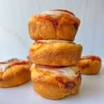 Three mini pizza muffins, made using a classic Pizza Bites Recipe, are stacked on a white plate with melted cheese and tomato sauce on top. A fourth muffin sits in the background against a clean, light backdrop.