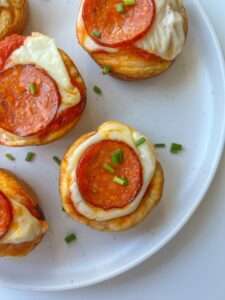 Mini pizza bites topped with melted cheese, pepperoni slices, and chopped green chives are arranged on a white plate—perfect for any gathering and inspired by a classic Pizza Bites Recipe.