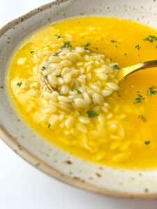 A close-up of Easy Pastina Soup, featuring a bowl filled with small pasta, possibly orzo, in a golden broth. A spoon lifts some pasta, and the comforting soup is garnished with chopped herbs.