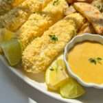 A plate of Fish and Chips featuring breaded fish fillets, potato wedges, lemon and lime slices, and a small bowl of yellow dipping sauce, all garnished with fresh herbs.