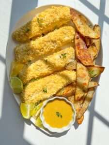 A plate of Fish and Chips featuring three breaded fish fillets, seasoned potato wedges, lime wedges, and a small dish of yellow dipping sauce, all garnished with fresh herbs and served in bright natural light.