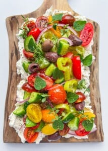A vibrant Greek Salad Feta Board features cherry tomatoes, cucumbers, olives, red onion, peppers, and fresh herbs atop whipped feta, beautifully arranged on a rustic wooden serving board.