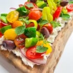 A wooden board topped with whipped cheese, fresh cherry tomatoes, cucumbers, olives, red onions, yellow peppers, and herbs—this Greek Salad Feta Board is a colorful and vibrant appetizer for any occasion.