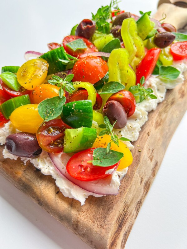 A wooden board topped with whipped cheese, fresh cherry tomatoes, cucumbers, olives, red onions, yellow peppers, and herbs—this Greek Salad Feta Board is a colorful and vibrant appetizer for any occasion.