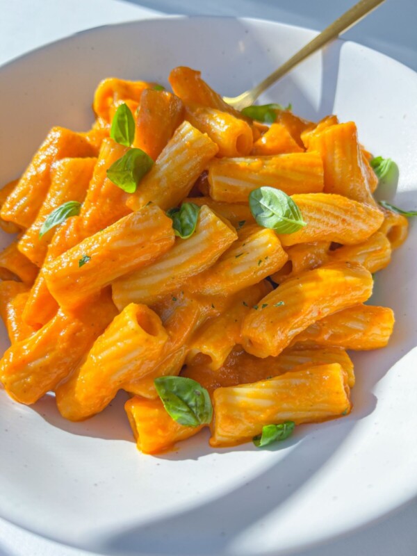 A white plate of rigatoni pasta coated in creamy orange Hidden Veggie Pasta Sauce, garnished with fresh basil leaves, with a gold fork resting on the side.