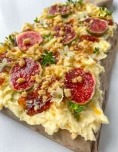 A close-up of whipped butter spread on a wooden board, topped with fresh fig slices, chopped nuts, honey, and garnished with small green herbs and white edible flowers—perfect inspiration if you’re wondering how to make a butter board.