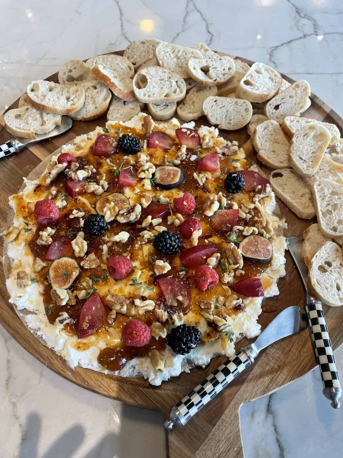 A wooden platter with whipped cheese topped with berries, figs, nuts, and herbs, surrounded by slices of baguette. Two cheese knives with black-and-white handles are placed next to the spread.