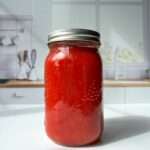 A glass jar filled with bright red Italian Tomato Sauce sits on a white countertop in a modern kitchen with white cabinets and a window in the background.