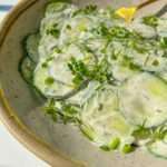 A bowl of creamy Polish Cucumber Salad garnished with fresh dill, green onions, and thyme, featuring thin cucumber slices coated in a white dressing and a yellow spoon resting in the bowl.
