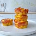 Four muffin-sized Pizza Egg Muffins are stacked on a white plate in a bright kitchen, with one egg bite resting beside the stack. This high protein breakfast recipe is perfect for busy mornings. The background is softly blurred.