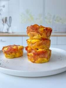 Four muffin-sized Pizza Egg Muffins are stacked on a white plate in a bright kitchen, with one egg bite resting beside the stack. This high protein breakfast recipe is perfect for busy mornings. The background is softly blurred.