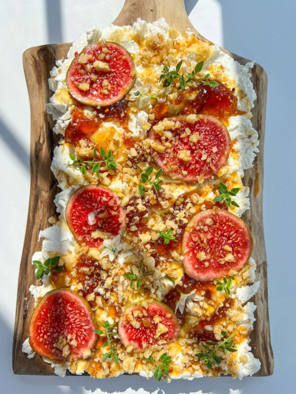A wooden board topped with whipped ricotta, fresh figs, chopped nuts, honey drizzle, and green herbs—an artistic take inspired by the viral goat cheese board trend, beautifully arranged on a white background.